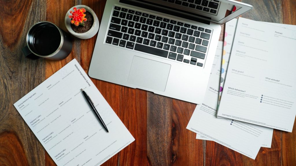 A photograph of a desktop covered in paperwork, and a laptop and coffee mug.